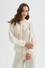 Blusa de algodón color beige, con cuello redondo y cierre frontal con botones. Presenta delicados bordados decorativos en tono tostado sobre el pecho y los hombros, mangas largas abullonadas con puños ajustados y diseño holgado.