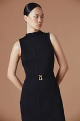 Vestido midi sin mangas, de cuello alto y textura plisada con ondas verticales. Presenta un corte entallado con un cinturón negro integrado que incluye una hebilla metálica dorada con el logo de la marca.