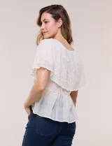 Blusa de estilo romántico color beige confeccionada en algodón, con escote amplio, importante volante capa en broderie calado, cartera frontal con botones tipo perla, bordados verticales calados, cintura marcada con bajo peplum y ruedo recto festonado.