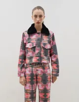 Chaqueta de denim con estampado abstracto en tonos rosados, naranjas y negros. Presenta un corte recto, cierre frontal con botones metálicos, bolsillos frontales con solapa y un cuello de piel sintética desmontable en color negro.
