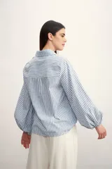 Camisa a rayas verticales color beige y blanco, con cuello clásico, cierre frontal con botón dorado, mangas largas abullonadas con puños elásticos, dos bolsillos frontales y corte holgado.