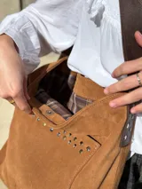 Cartera tipo tote bag de cuero color camel, con detalles de tachas metálicas en el bolsillo frontal y tiras largas para el hombro.