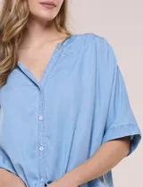 Blusa estilo denim de lyocell celeste, de silueta amplia y suelta, con escote en V, cartera frontal de botones y mangas cortas japonesas de hombros caídos. Cuenta con ruedo asimétrico y posibilidad de anudar al frente.