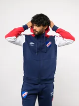 Campera deportiva Umbro del Club Nacional de Football, con cierre frontal, capucha ajustable con cordones y diseño en bloques de color azul, celeste y blanco. Presenta el logo de la marca y el escudo del club bordados en el pecho.