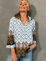 Blusa de manga larga con cuello camisero, corte recto y cierre frontal de botones. Presenta un diseño estampado geométrico en tonos celeste, blanco y marrón.