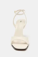 Sandalia de tacón alto en color blanco marfil, con tira delantera ancha, diseño destalonado y tira ajustable al tobillo con hebilla metálica dorada. Presenta punta cuadrada y tacón fino.