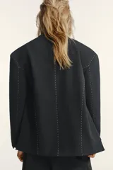 Blazer oversize de corte recto con cuello de solapa, manga larga con hombreras y cierre frontal de doble botonadura. Presenta un diseño en color gris oscuro con costuras decorativas a contraste en color blanco, bolsillos con solapa y forro interior.