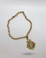 Collar de cadena de eslabones gruesos en acero dorado, con un dije central de diseño circular tipo moneda antigua con textura rugosa y una piedra rectangular clara engastada en el centro.