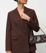 Blazer de sastrería color marrón, de corte regular y largo, con cuello peaked, cierre frontal cruzado con botones, bolsillos con tapa y mangas largas con botones decorativos.