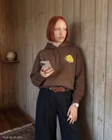 Canguro oversize color marrón con capucha y bolsillo frontal. Presenta un estampado gráfico en la espalda que incluye una ilustración de una mujer, texto decorativo y detalles alusivos a yerba mate.