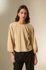 Blusa de color beige con cuello redondo y mangas largas abullonadas con puños elásticos. Presenta un corte holgado y fluido.