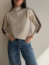 Sweater de tejido de punto en color beige con cuello alto y costura central vertical. Presenta mangas ranglan con una franja lateral en color marrón oscuro que genera contraste, y terminaciones en tejido acanalado en puños y ruedo.