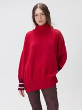 Pullover de punto color rojo, con cuello tortuga, corte holgado y mangas largas con puños acanalados en contraste con rayas blancas. Presenta un logo bordado en el pecho.