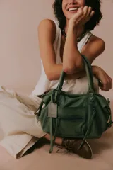 Cartera de cuero verde militar con acabado gastado, cuerpo blando, asas de mano, bolsillos frontales con cierre y tiras colgantes.
