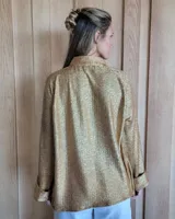 Camisa de corte holgado en color dorado con acabado brillante y textura plisada. Presenta cuello clásico, cierre frontal con botones y mangas largas con puños anchos doblados.