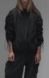 Campera rompevientos negra de calce oversize, confeccionada en microfibra técnica ligera con acabado semi-mate. Presenta capucha integrada, cierre frontal técnico y bolsillos tipo ojal. Destaca por su sistema de frunces elásticos regulables mediante tancas a lo largo de las mangas y el ruedo, permitiendo ajustar el volumen de la prenda.