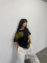 Remera negra de corte regular con estampado gráfico en color amarillo. Presenta inscripciones con estética oriental en la parte superior izquierda y en el lateral derecho. Confeccionada en algodón de alto gramaje con cuello reforzado y grifa de la marca en la manga.