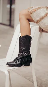 Bota de cuero negro estilo texana con caña media. Presenta un aplique desmontable en la zona del tobillo decorado con tachas metálicas y flecos largos. Posee taco bajo y punta levemente afinada.