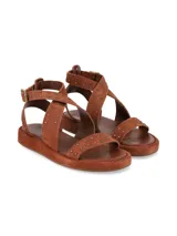 Sandalia plana de gamuza marrón chocolate, con tiras cruzadas decoradas con tachas metálicas doradas y tira ajustable al tobillo con hebilla metálica.