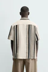 Camisa de manga corta con calce relaxed fit, confeccionada en tejido estructurado con algodón. Presenta un diseño de rayas verticales en tonos beige, negro y blanco, cuello solapa, cierre frontal con botones y aberturas laterales en el bajo.