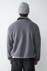 Campera de tejido polar tipo teddy en color gris, con cuello camisero, cierre frontal de cremallera y bolsillo funcional en el pecho con solapa de contraste.