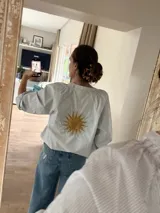Camisa de corte suelto con estampado de rayas finas, cuello redondo y mangas largas con puños elásticos. Presenta un bordado central en la espalda con diseño de sol en color amarillo.