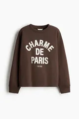 Buzo oversize de color marrón, con cuello redondo, mangas largas de hombros caídos y estampa frontal en color blanco con la leyenda "Charme de Paris 1998".