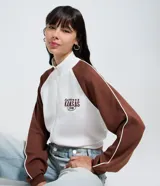 Saco femenino de poliviscosa, color blanco con mangas raglán marrones y ribetes blancos. Tiene cuello alto, cierre frontal de cremallera y bordado lettering "Kansas City" en el pecho.