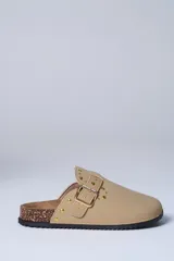 Zueco de gamuza sintética color beige, con detalle de tachas metálicas doradas y hebilla lateral ajustable. Presenta una base de corcho y suela de goma dentada.