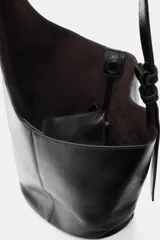 Bolso tipo saca de diseño asimétrico confeccionado en piel negra con acabado brillante. Cuenta con asa de hombro ajustable y cierre mediante imán. Incluye un bolsillo extraíble en su interior.