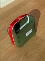 Necessaire de neopreno color verde oliva, con cierre superior y manija de cinta en color rojo y beige. Presenta un aplique frontal con el logo de la marca.