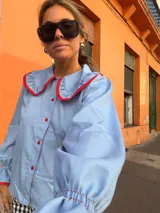Camisa de color celeste con cuello bobo grande y puños con volados, ambos con ribetes en color rojo. Presenta cierre frontal con botones rojos.
