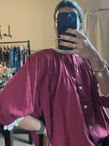 Camisa bordó de corte holgado y diseño abullonado, con cuello redondo alto y puños adornados con detalles de encaje blanco. Presenta pliegues frontales y cierre con botones nacarados.