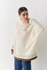 Buzo tejido de punto con cuello alto, diseño holgado y aberturas laterales. Presenta un cuerpo en color azul melange con detalles de franjas en color crema y verde en los puños y el ruedo. Incluye un pequeño bordado en el pecho.