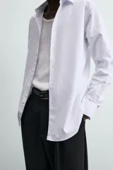 Camisa slim fit blanca con rayas verticales finas en color gris, confeccionada en tejido de algodón. Presenta cuello italiano y manga larga con puño abotonado. Cierre frontal con botonadura.
