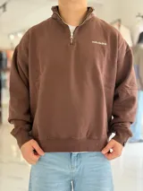 Buzo gris oscuro de corte oversize con cuello alto y cierre frontal parcial. Cuenta con mangas largas y puños elásticos, además de un pequeño bordado con texto en el pecho.