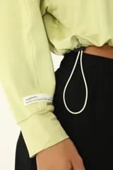 Buzo crop color verde claro, con cuello redondo y mangas largas holgadas. Presenta frunce en el ruedo mediante cordón elástico y tanca.