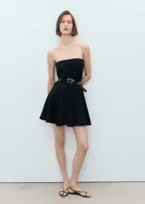 Vestido corto negro con escote palabra de honor, diseño evasé y falda con tablas. Cuenta con cierre de cremallera en la parte posterior.