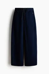 Jean de denim rígido azul oscuro, con corte holgado y diseño curvo en las piernas. Presenta cintura estándar con cordón ajustable, cierre con botón y bolsillos clásicos.