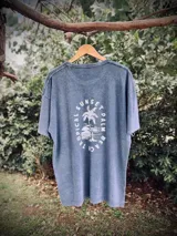 Remera oversize de algodón con efecto lavado (acid wash) en tono azul grisáceo. Presenta un estampado circular en la espalda con una palmera y texto que dice "SUNSET", "TROPICAL" y "PALM BEACH".