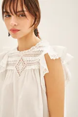 Camisa blanca de algodón con detalles de puntilla en el canesú, volados con terminación de puntillas en las mangas y cartera de botones en la parte superior de la espalda.