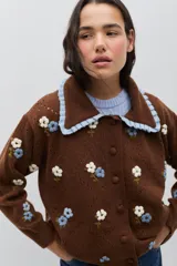 Saco de punto color marrón con bordados artesanales de flores en tonos blanco y celeste. Presenta un cuello con volados en color celeste, cierre frontal con botones y diseño de calados sutiles en el tejido.