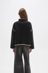 Polera de tejido de punto color negro con detalles de vivos en color beige en el cuello, puños, ruedo y costuras de las mangas. Presenta un diseño holgado con cuello alto y mangas largas.