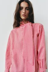 Camisa de rayas rosadas y blancas, con cuello subido decorado con un lazo integrado, manga larga con puños abotonados y cierre frontal oculto.