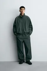 Chaqueta técnica de corte relajado en color verde oscuro, con cuello subido, cierre frontal de cremallera y detalles de líneas en contraste. Presenta bolsillos frontales con cremallera oculta y acabados elásticos en puños y ruedo.