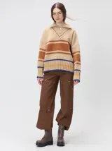 Pullover de punto con textura aterciopelada, diseño oversize, cuello polo con cierre frontal parcial y detalles de pespuntes en contraste. Presenta rayas horizontales en tonos tierra y un parche bordado con la letra M en el pecho.