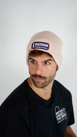 Gorro tipo beanie de punto acanalado en color beige, con parche rectangular bordado en el frente que incluye el logo y nombre de la marca Duxon.