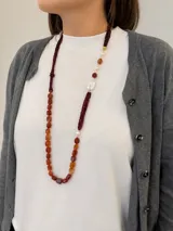 Collar largo de diseño asimétrico compuesto por múltiples tiras finas de abalorios bordó, combinado con piedras naturales de cornalina en tonos rojizos y naranjas, perlas barrocas blancas y detalles metálicos dorados.