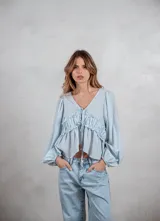 Camisa de tela liviana color celeste, con escote en V y cierre frontal mediante lazos ajustables. Presenta un diseño con volados horizontales en el pecho y mangas largas abullonadas con puños elásticos.