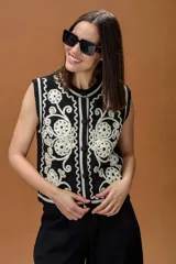 Chaleco tejido sin mangas, color negro con detalles bordados en color crema con diseño de arabescos y flores. Presenta cierre frontal con botones y cuello redondo.